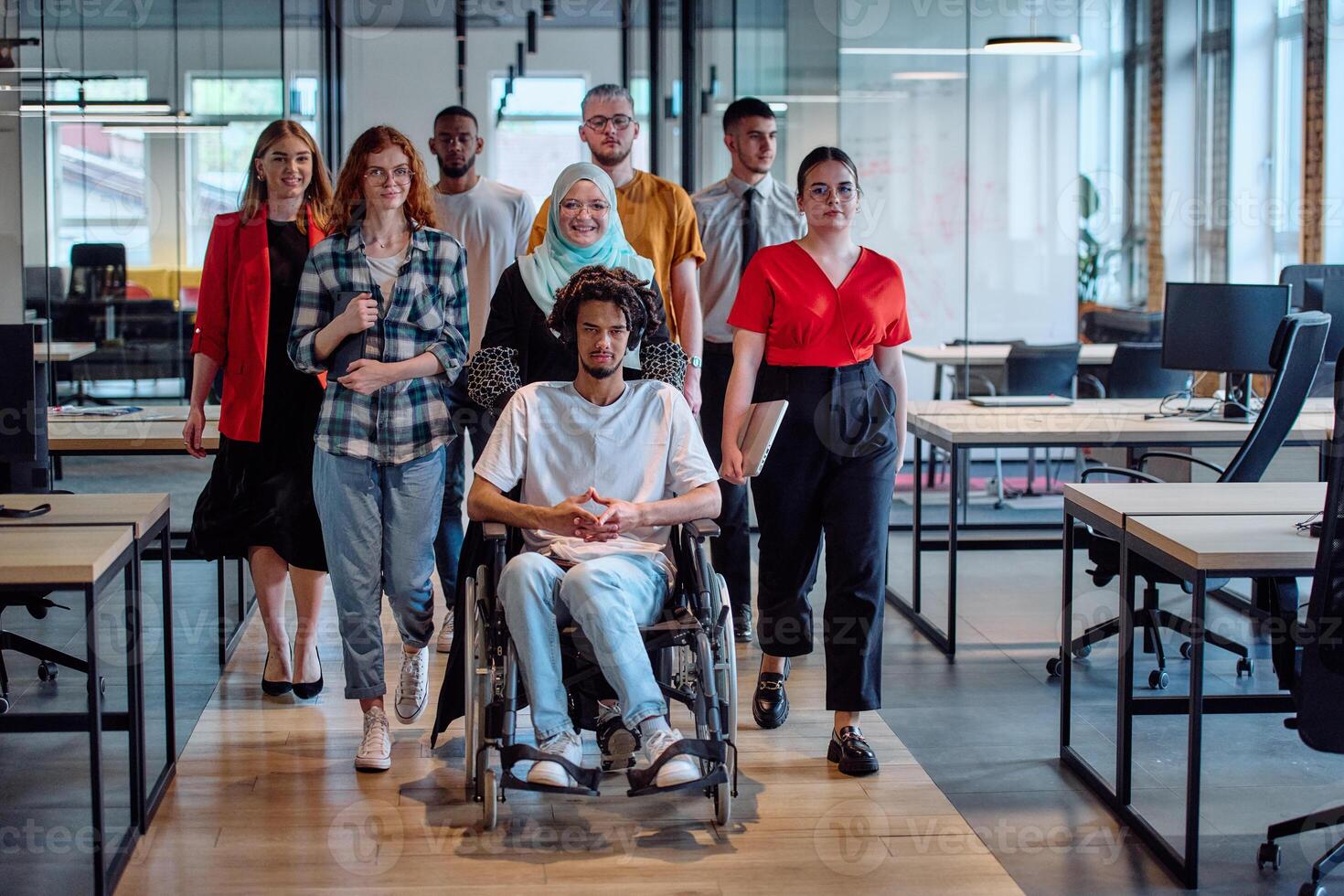 A diverse group of young business people congregates within a modern startup's glass-enclosed office, featuring inclusivity with a person in a wheelchair, an African American young man, and a hijab photo