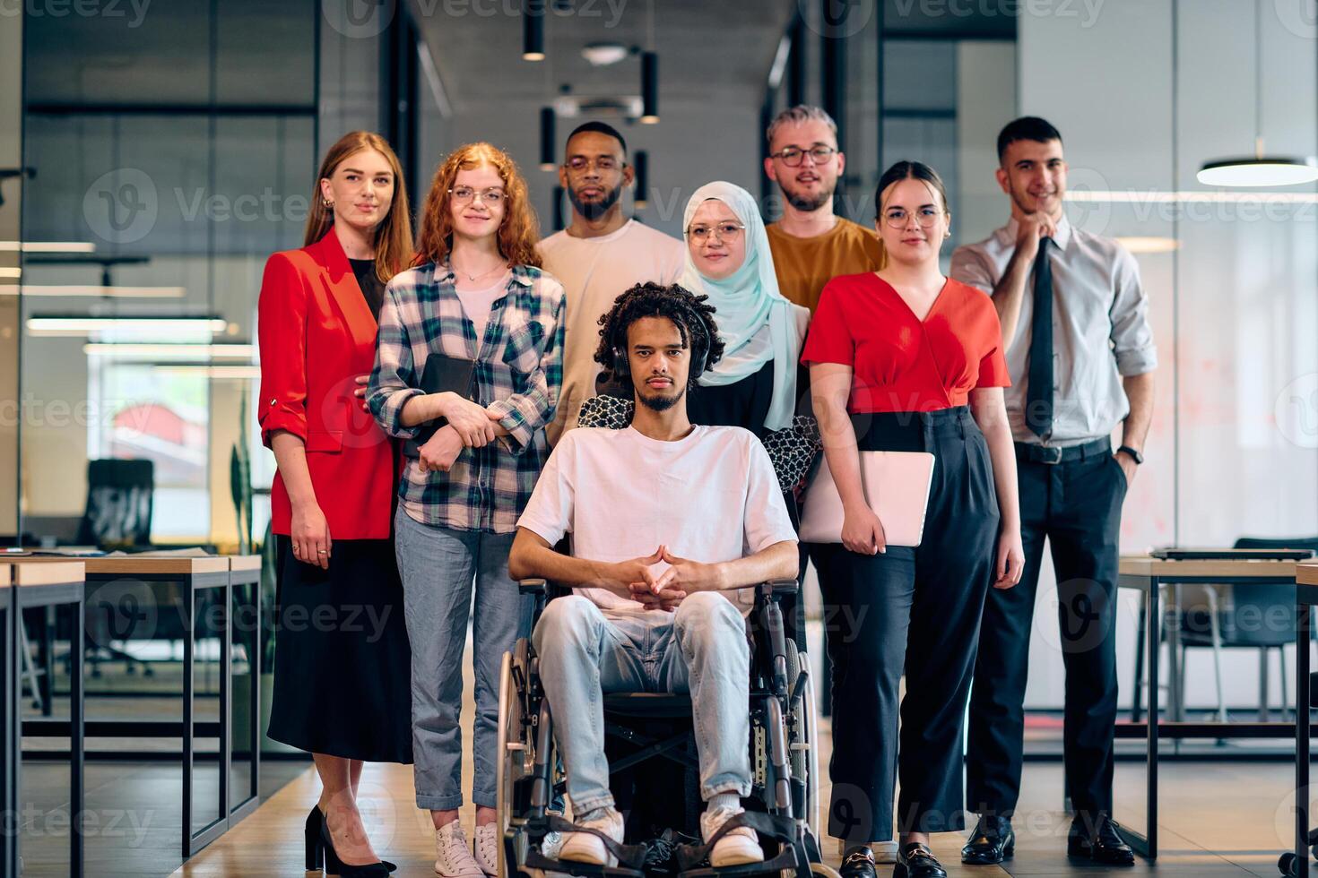A diverse group of young business people walking a corridor in the glass-enclosed office of a modern startup, including a person in a wheelchair and a woman wearing a hijab photo