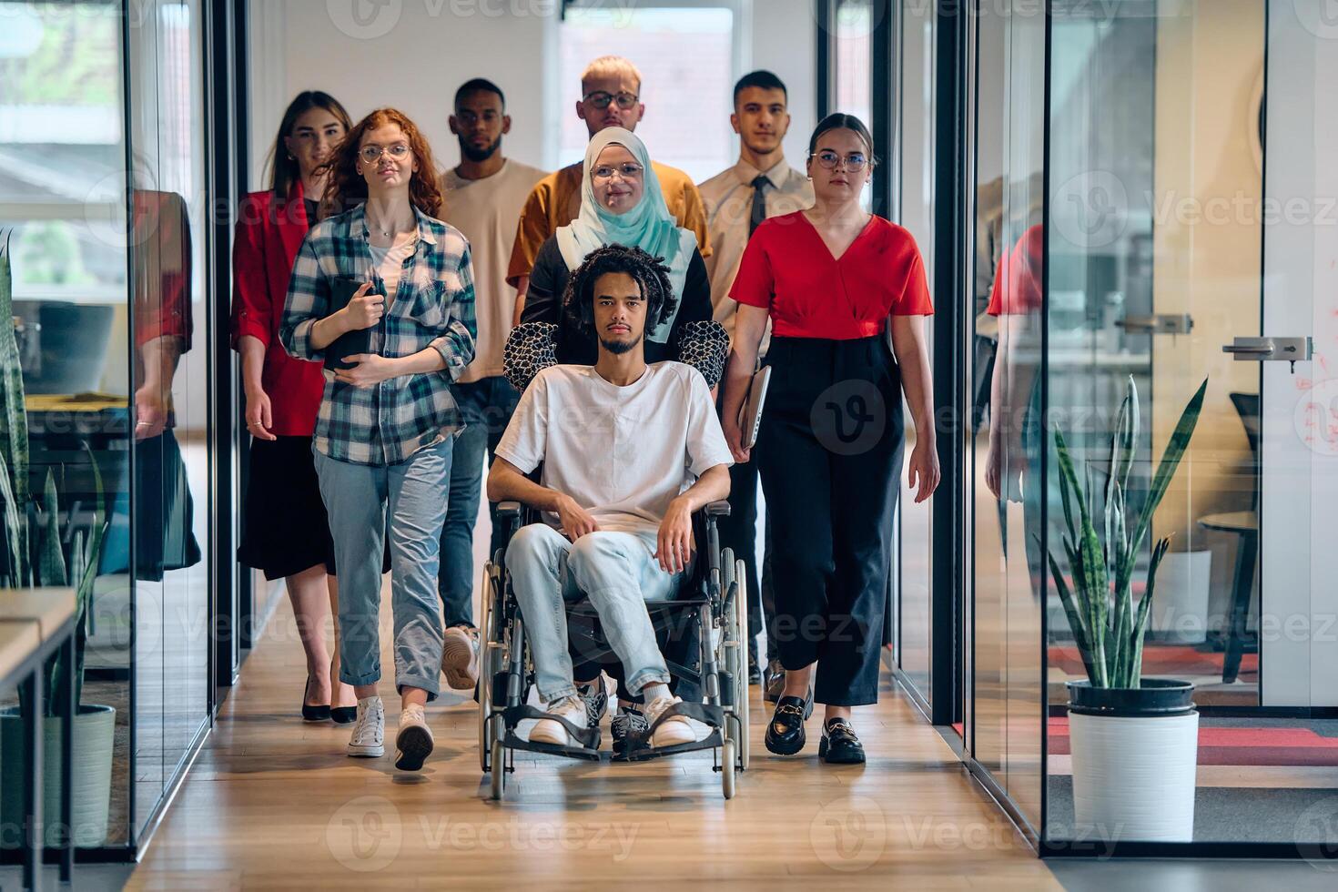 A diverse group of young business people congregates within a modern startup's glass-enclosed office, featuring inclusivity with a person in a wheelchair, an African American young man, and a hijab photo