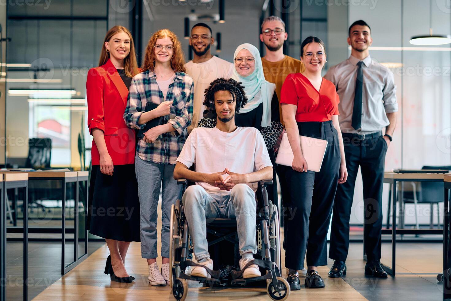 A diverse group of young business people walking a corridor in the glass-enclosed office of a modern startup, including a person in a wheelchair and a woman wearing a hijab photo