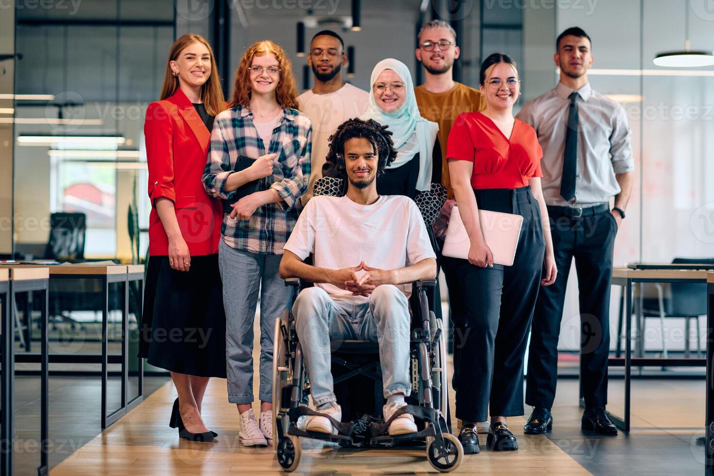 A diverse group of young business people walking a corridor in the glass-enclosed office of a modern startup, including a person in a wheelchair and a woman wearing a hijab photo