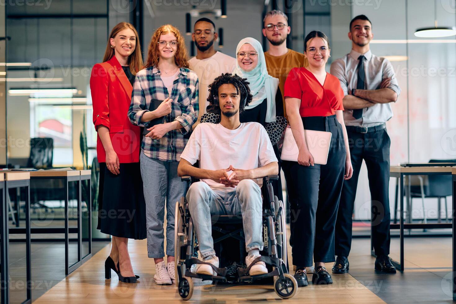 A diverse group of young business people walking a corridor in the glass-enclosed office of a modern startup, including a person in a wheelchair and a woman wearing a hijab photo
