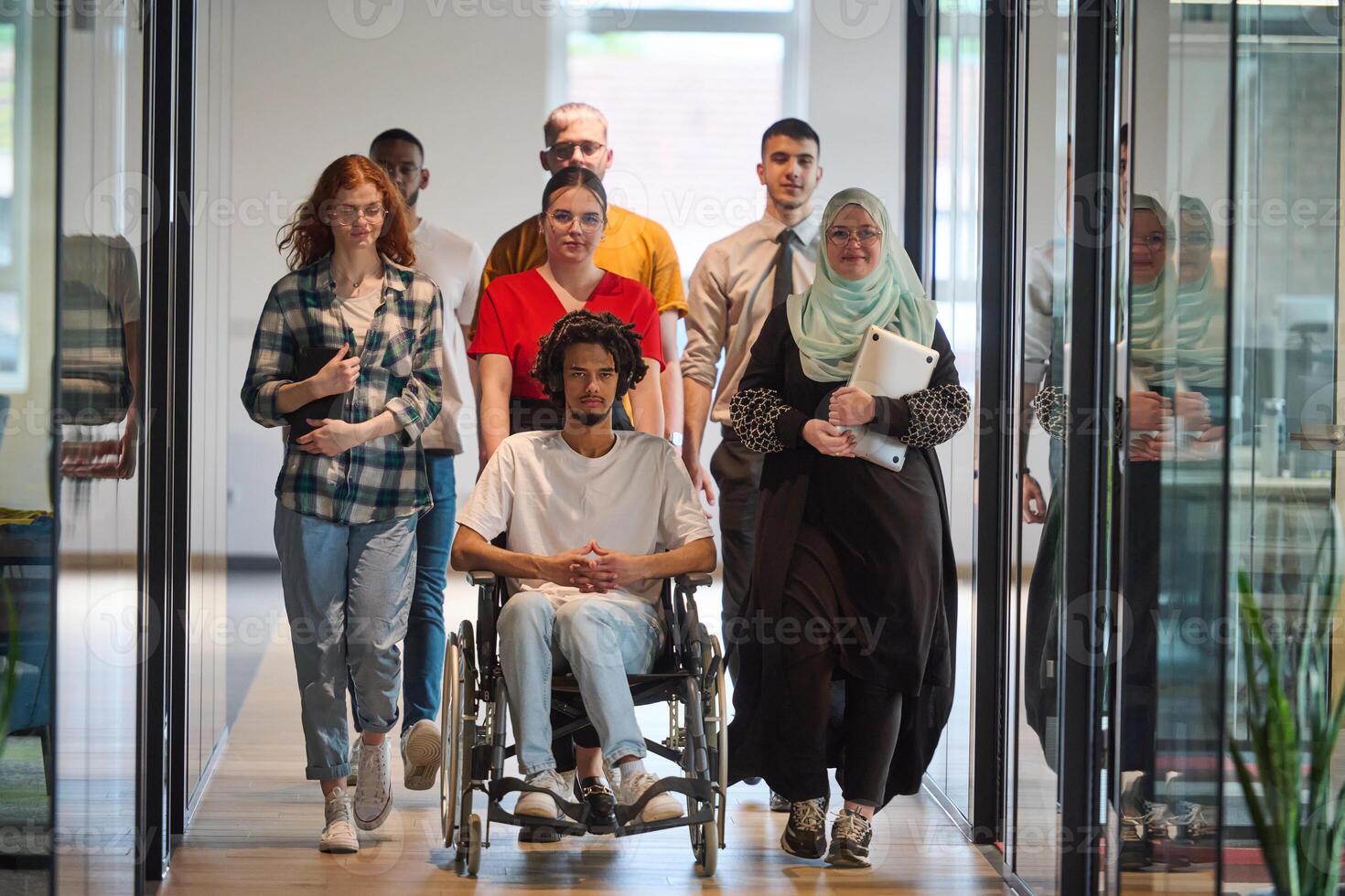 A diverse group of young business people congregates within a modern startup's glass-enclosed office, featuring inclusivity with a person in a wheelchair, an African American young man, and a hijab photo