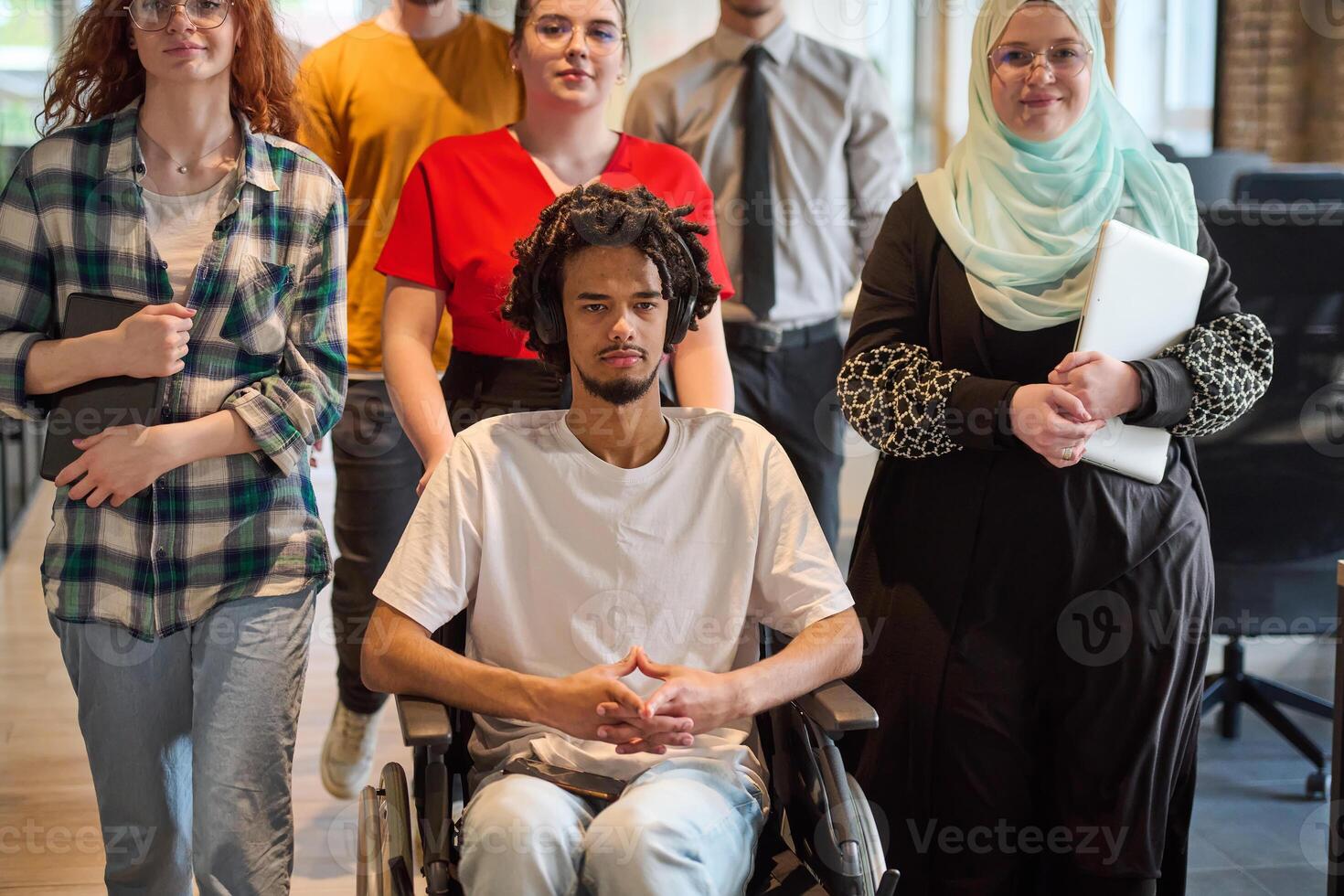 A diverse group of young business people congregates within a modern startup's glass-enclosed office, featuring inclusivity with a person in a wheelchair, an African American young man, and a hijab photo