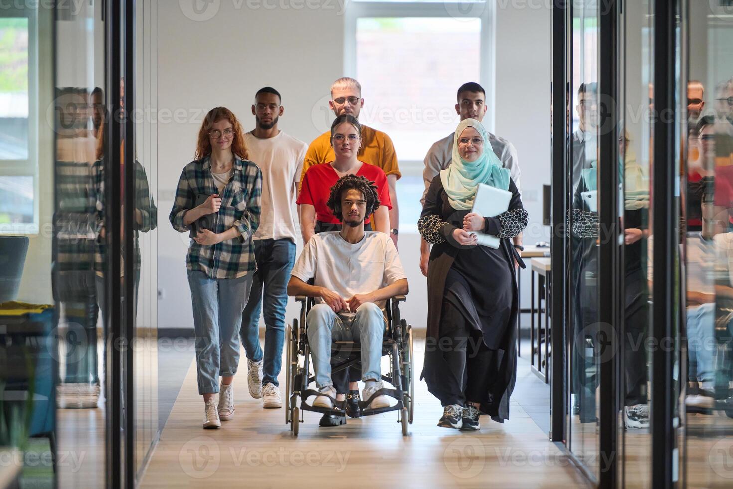 A diverse group of young business people congregates within a modern startup's glass-enclosed office, featuring inclusivity with a person in a wheelchair, an African American young man, and a hijab photo