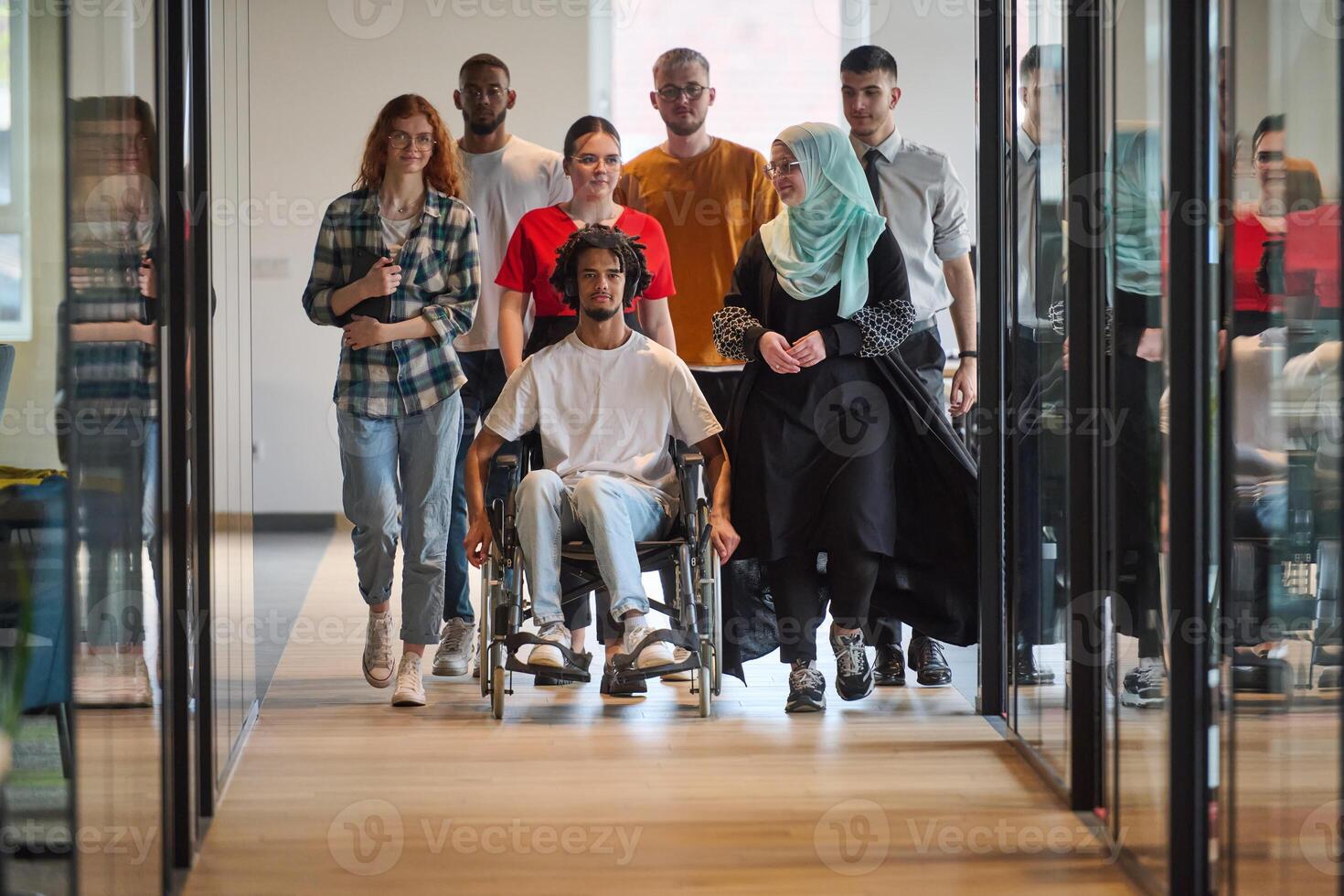 A diverse group of young business people congregates within a modern startup's glass-enclosed office, featuring inclusivity with a person in a wheelchair, an African American young man, and a hijab photo
