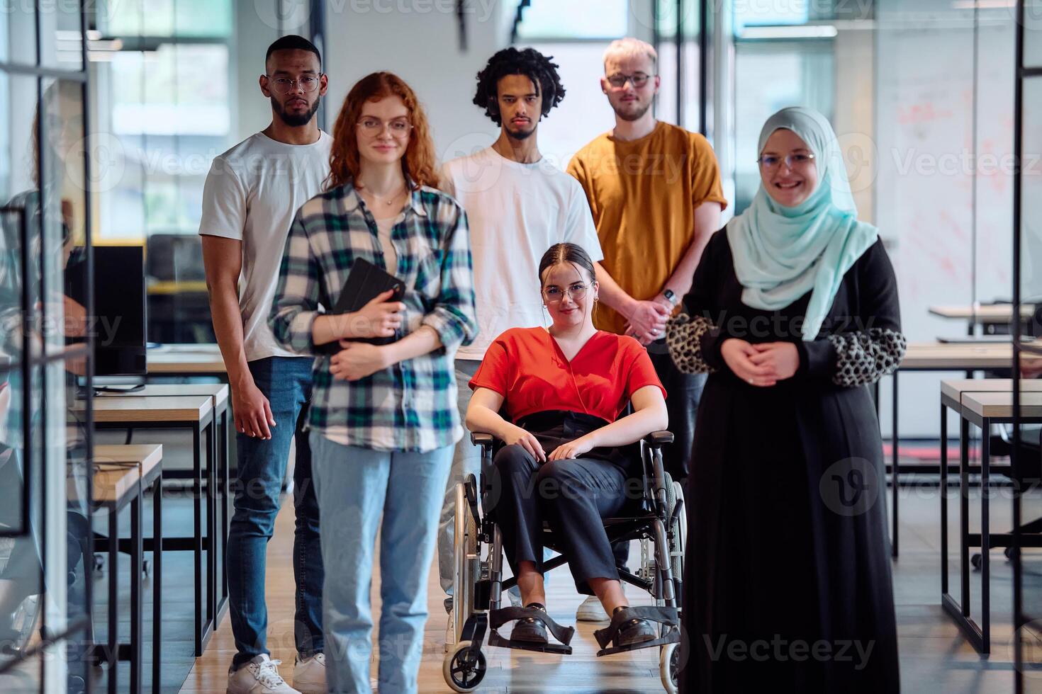 A diverse group of young business people walking a corridor in the glass-enclosed office of a modern startup, including a person in a wheelchair and a woman wearing a hijab photo