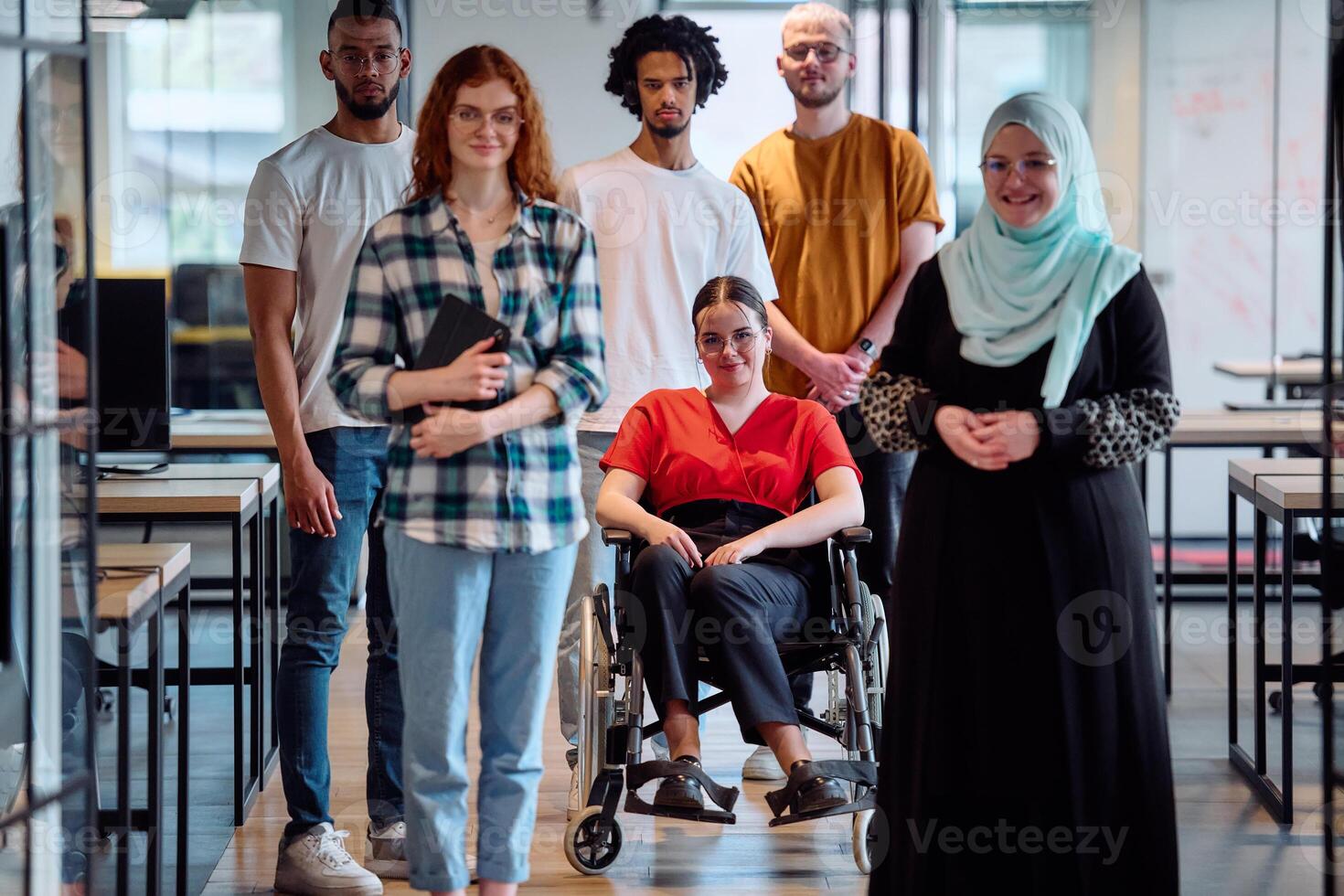A diverse group of young business people walking a corridor in the glass-enclosed office of a modern startup, including a person in a wheelchair and a woman wearing a hijab photo