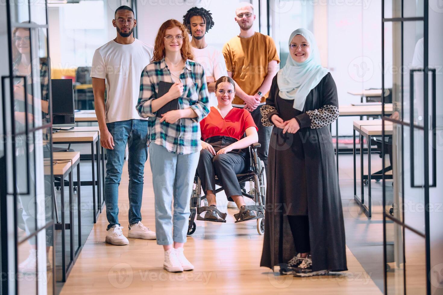 A diverse group of young business people walking a corridor in the glass-enclosed office of a modern startup, including a person in a wheelchair and a woman wearing a hijab photo