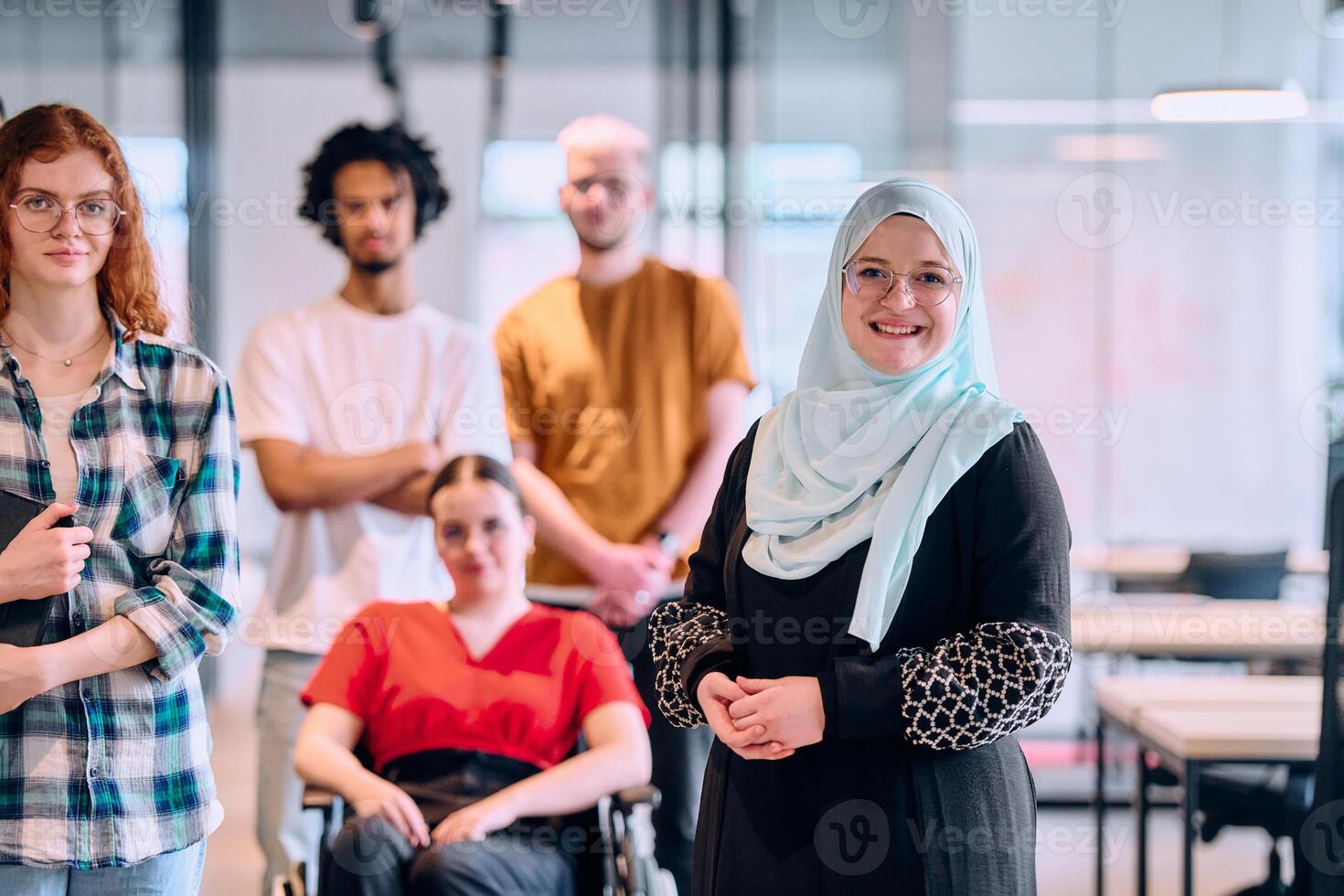 A diverse group of young business people walking a corridor in the glass-enclosed office of a modern startup, including a person in a wheelchair and a woman wearing a hijab photo