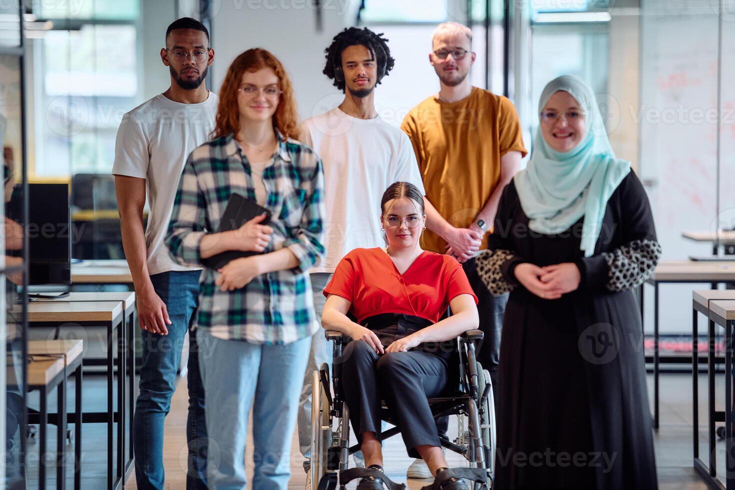 A diverse group of young business people walking a corridor in the glass-enclosed office of a modern startup, including a person in a wheelchair and a woman wearing a hijab photo