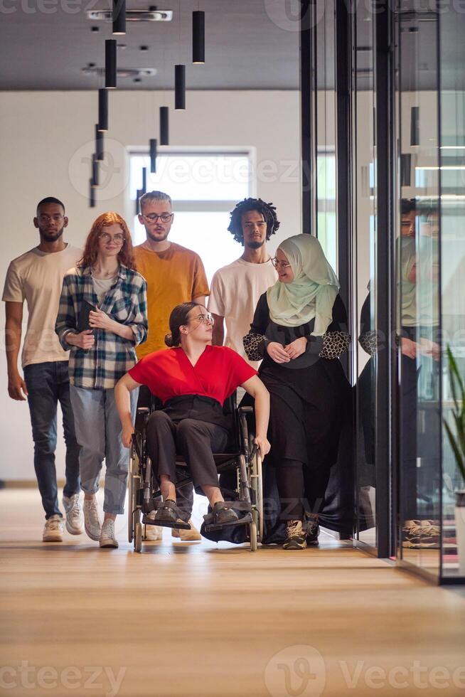 A diverse group of young business people walking a corridor in the glass-enclosed office of a modern startup, including a person in a wheelchair and a woman wearing a hijab photo
