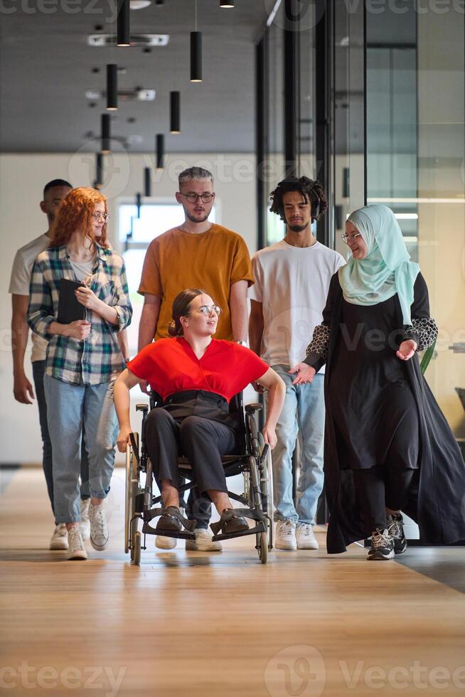 A diverse group of young business people walking a corridor in the glass-enclosed office of a modern startup, including a person in a wheelchair and a woman wearing a hijab photo