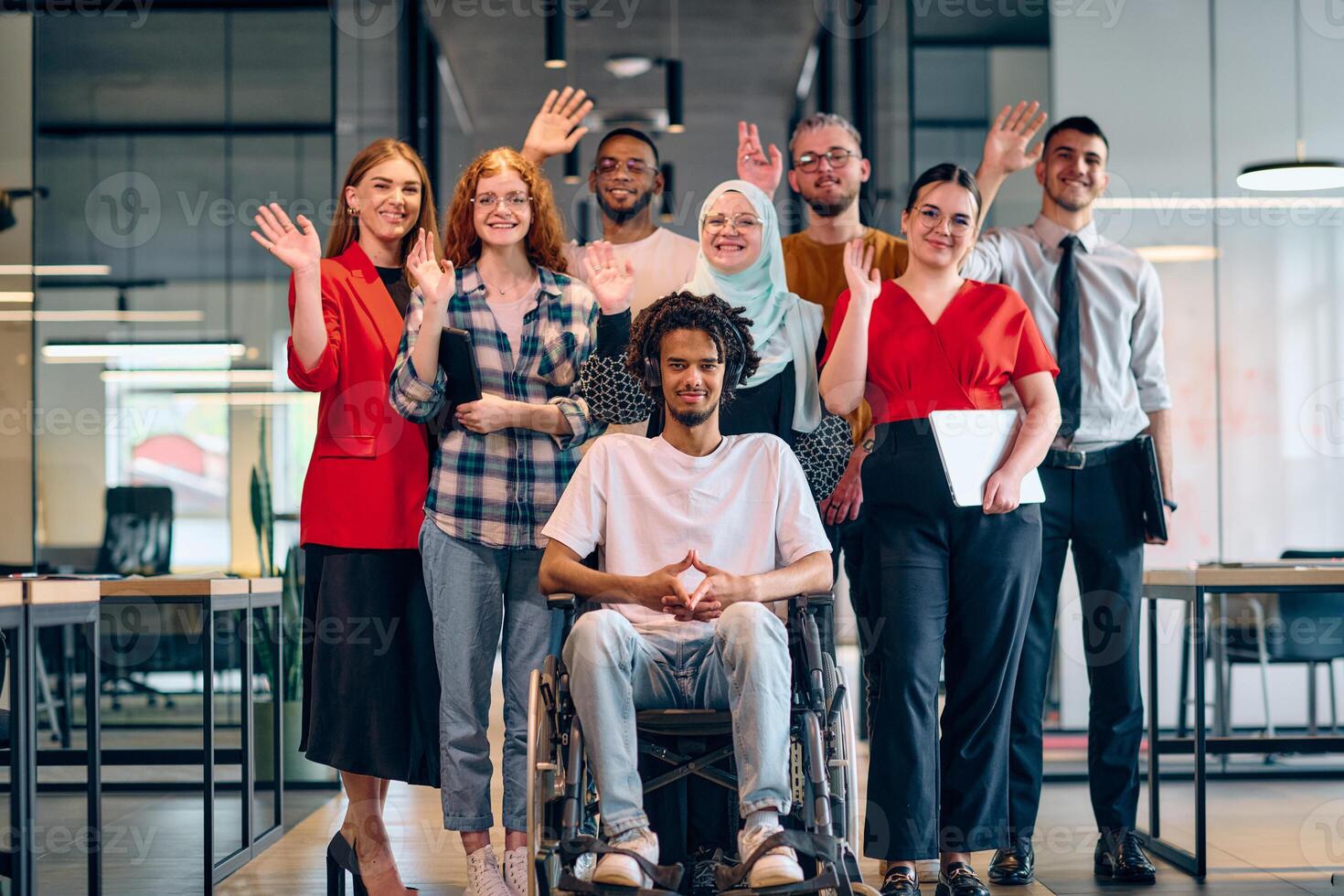A diverse group of young business people walking a corridor in the glass-enclosed office of a modern startup, including a person in a wheelchair and a woman wearing a hijab photo