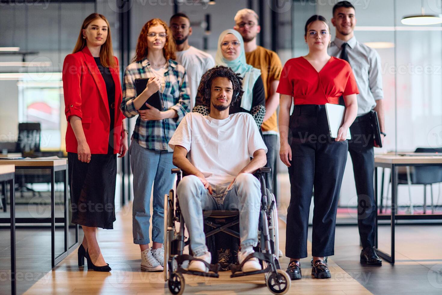 A diverse group of young business people walking a corridor in the glass-enclosed office of a modern startup, including a person in a wheelchair and a woman wearing a hijab photo