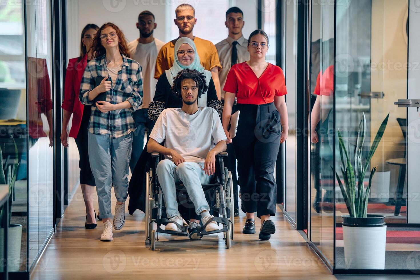 A diverse group of young business people congregates within a modern startup's glass-enclosed office, featuring inclusivity with a person in a wheelchair, an African American young man, and a hijab photo