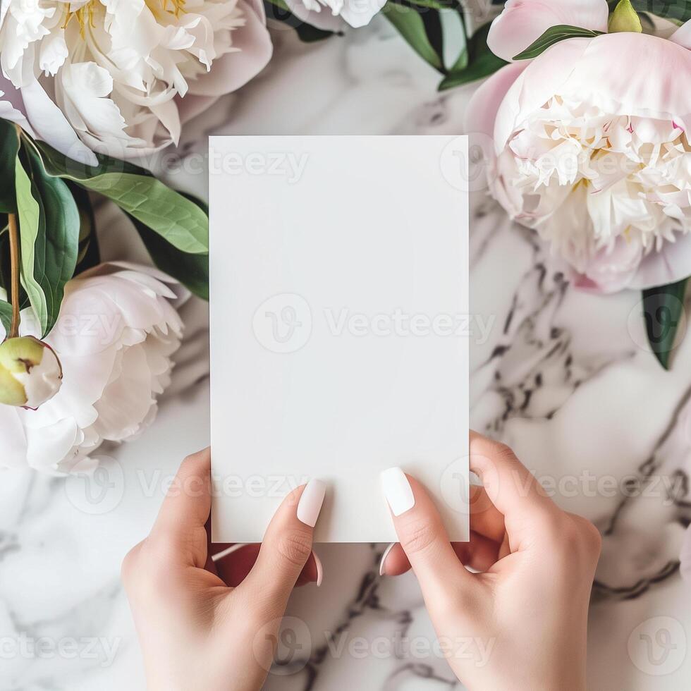 A clean and sophisticated mockup featuring a woman's hands holding a blank invitation card, surrounded by lush peonies and elegant decor on a marble background. photo