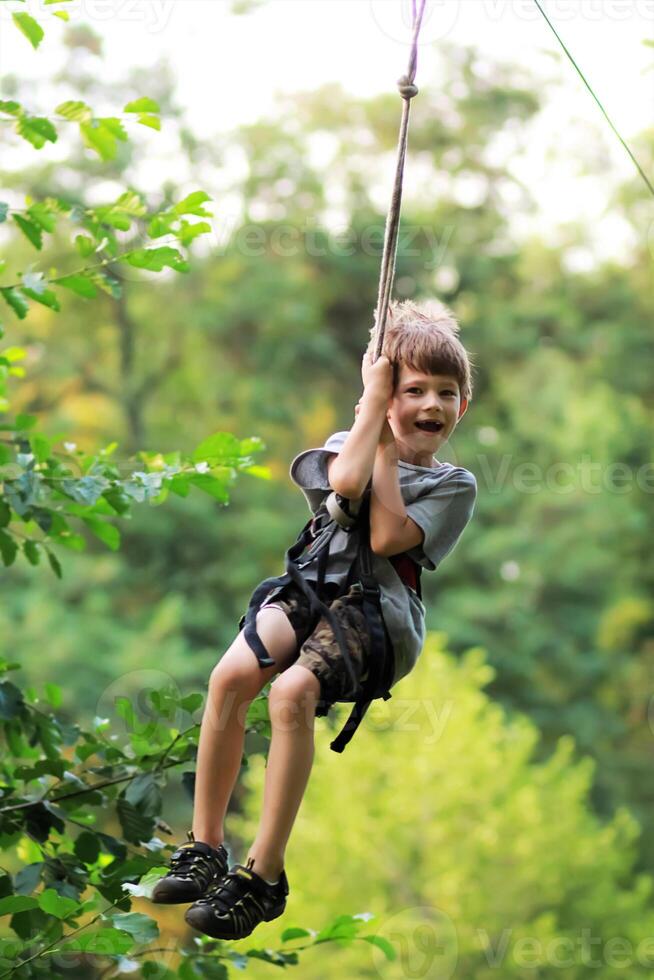 Happy shouting boy flying in the air on zip line in grey t shirt and shorts in the summer park among trees and lush foliage photo
