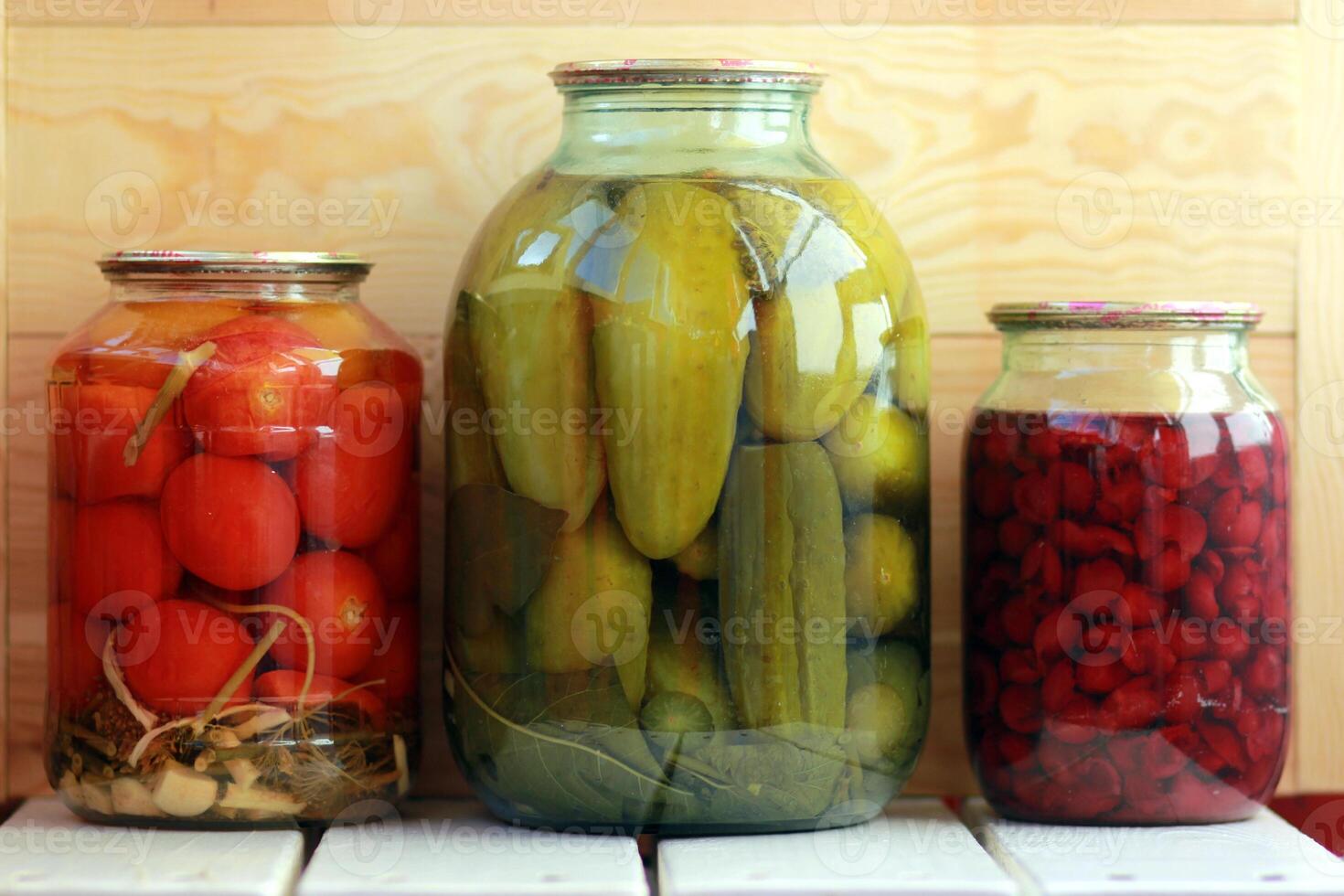 Three jars of pickled cucumbers, tomatoes and cherry jam against the wooden background photo