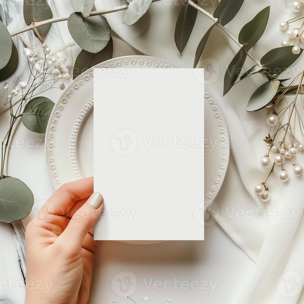 Blank white paper card mockup on a plate with woman's hand holding it. Styled with eucalyptus leaves and baby's breath flowers. photo