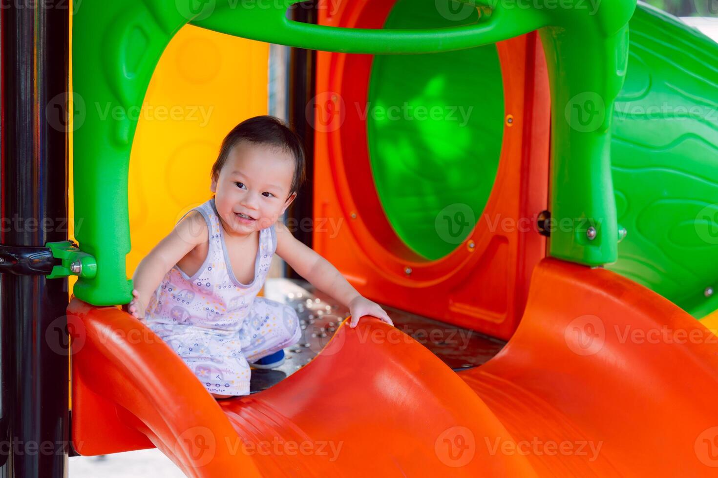 Portrait of A happy toddler boy playing on a playground orange slide, in the summer or spring, enjoying on sunny day. Child aged one year old. photo