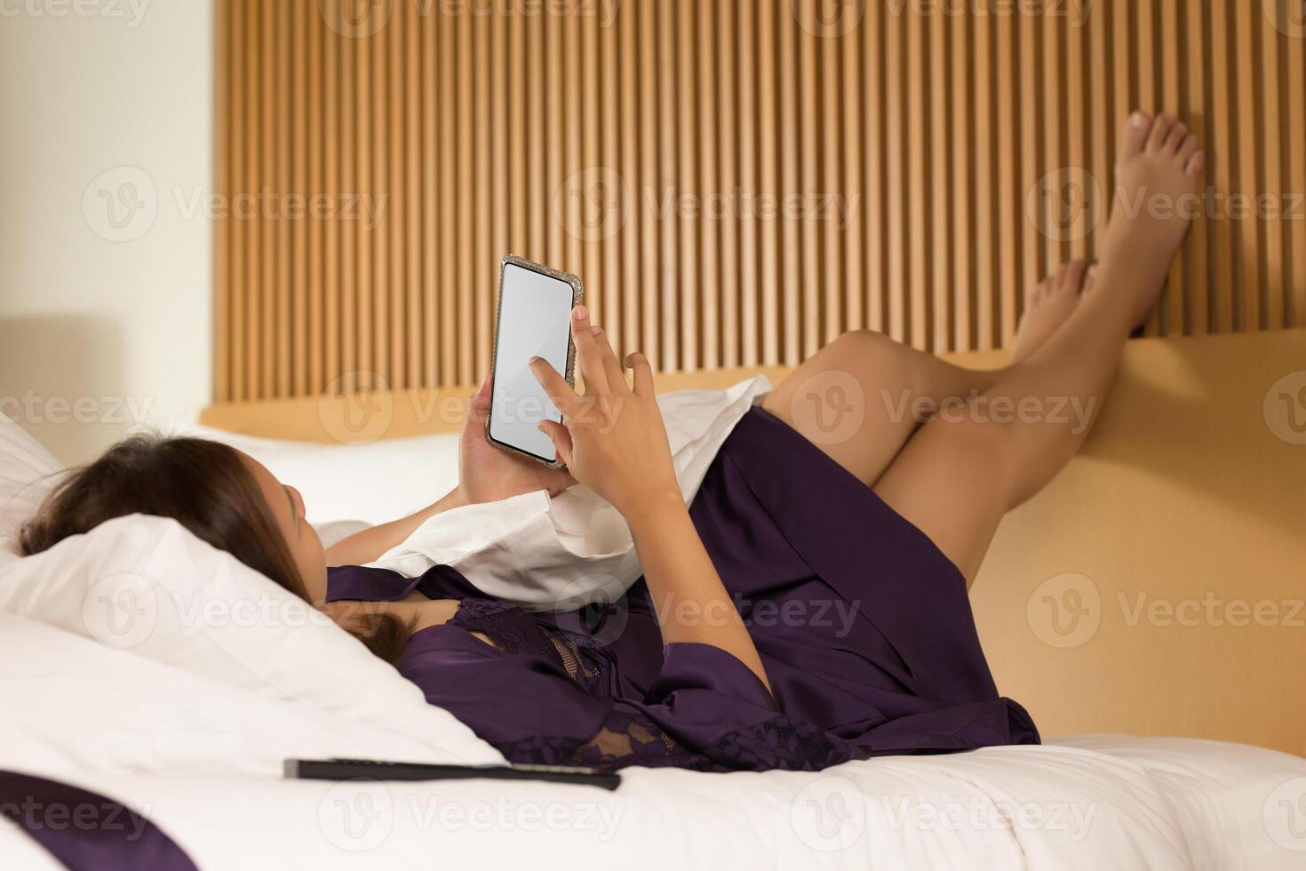 A woman, clad in a purple satin nightgown, lies supine on her bed, utilizing her smartphone during the nocturnal hours. photo