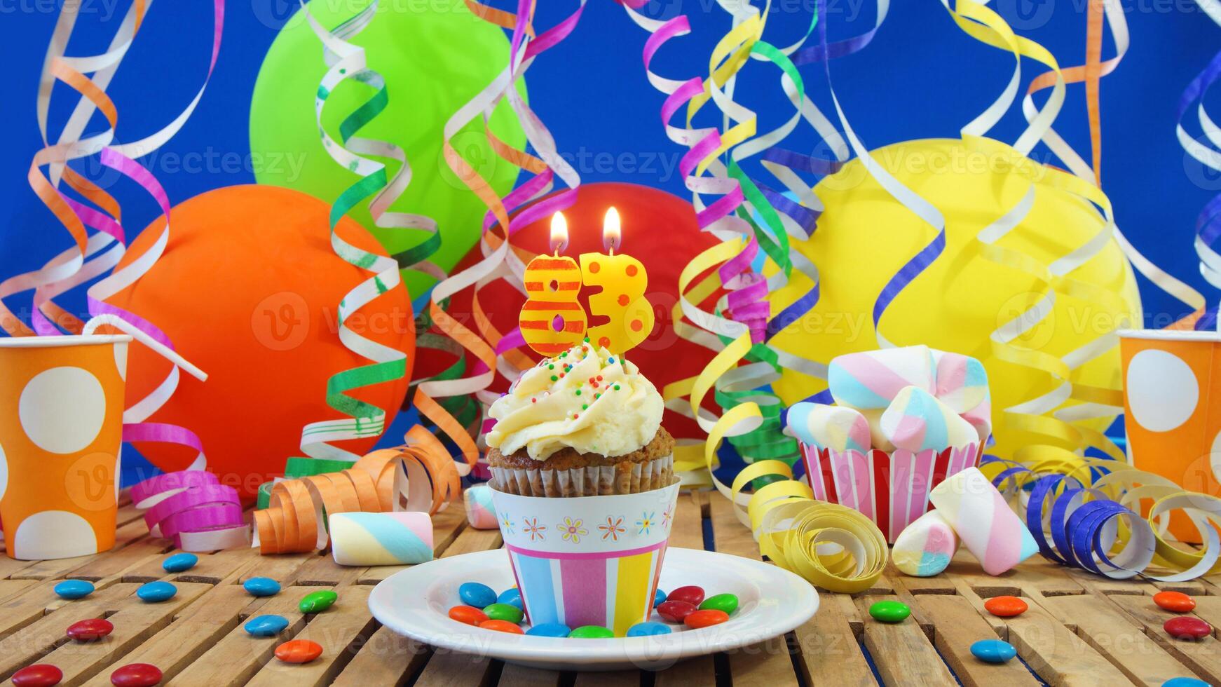 Birthday 83 cupcake with candles burning on rustic wooden table with background of colorful balloons, plastic cups and candies with blue wall in the background photo