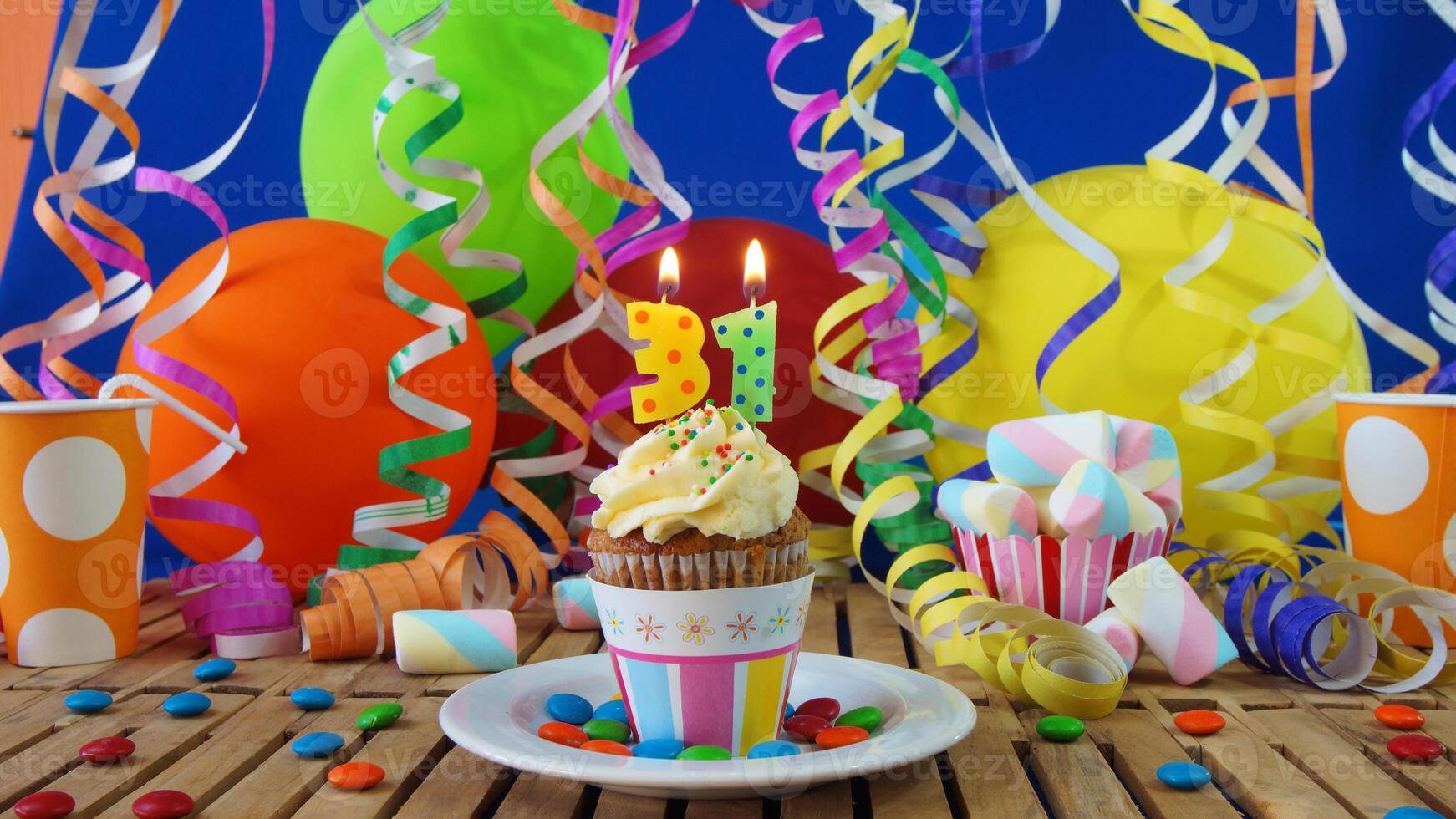 Birthday 31 cupcake with candles burning on rustic wooden table with background of colorful balloons, plastic cups and candies with blue wall in the background photo
