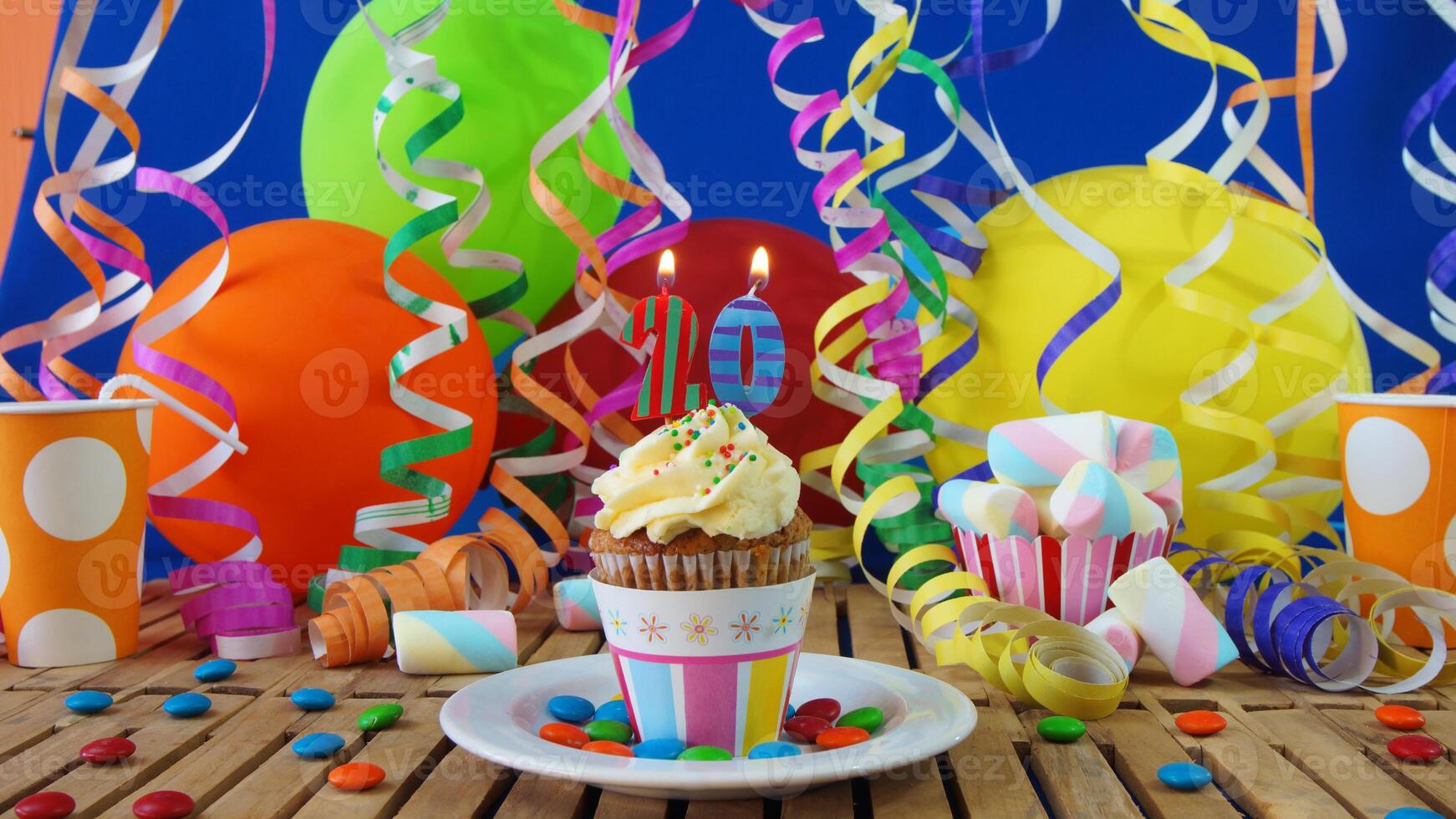 Birthday 20 cupcake with candles burning on rustic wooden table with background of colorful balloons, plastic cups and candies with blue wall in the background photo