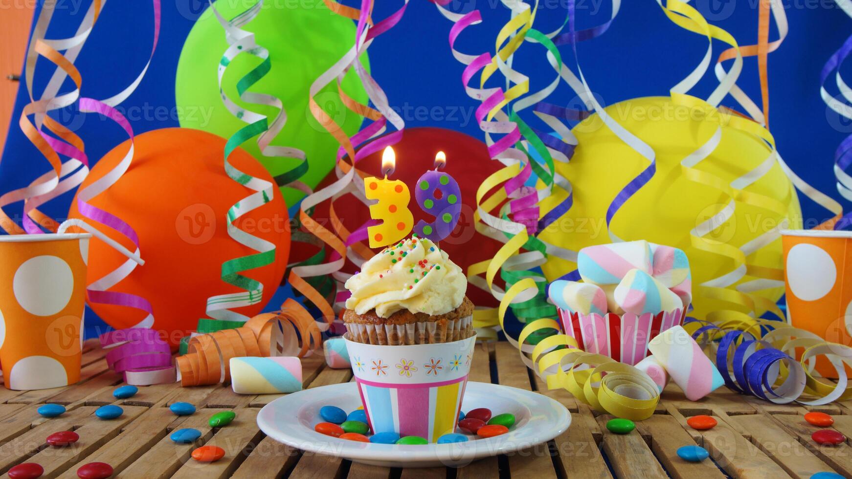 Birthday 39 cupcake with candles burning on rustic wooden table with background of colorful balloons, plastic cups and candies with blue wall in the background photo