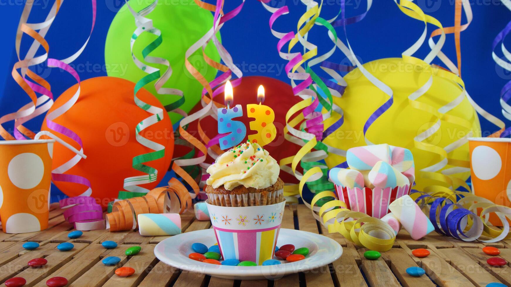 Birthday 53 cupcake with candles burning on rustic wooden table with background of colorful balloons, plastic cups and candies with blue wall in the background photo