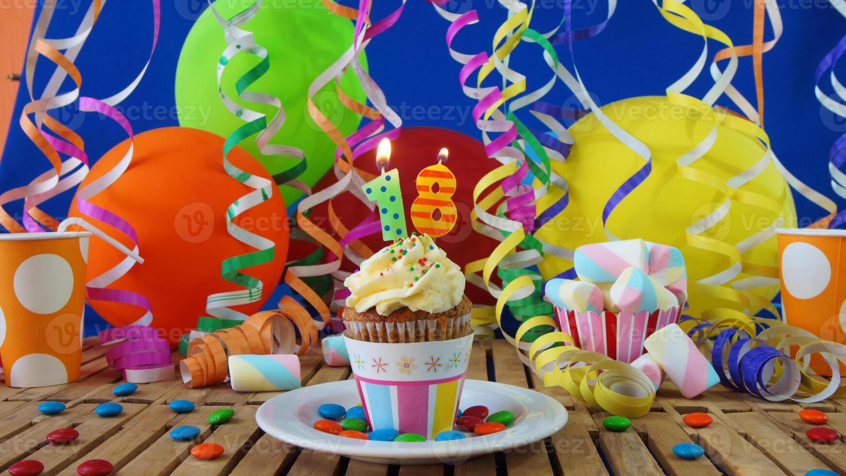 Birthday 18 cupcake with candles burning on rustic wooden table with background of colorful balloons, plastic cups and candies with blue wall in the background photo