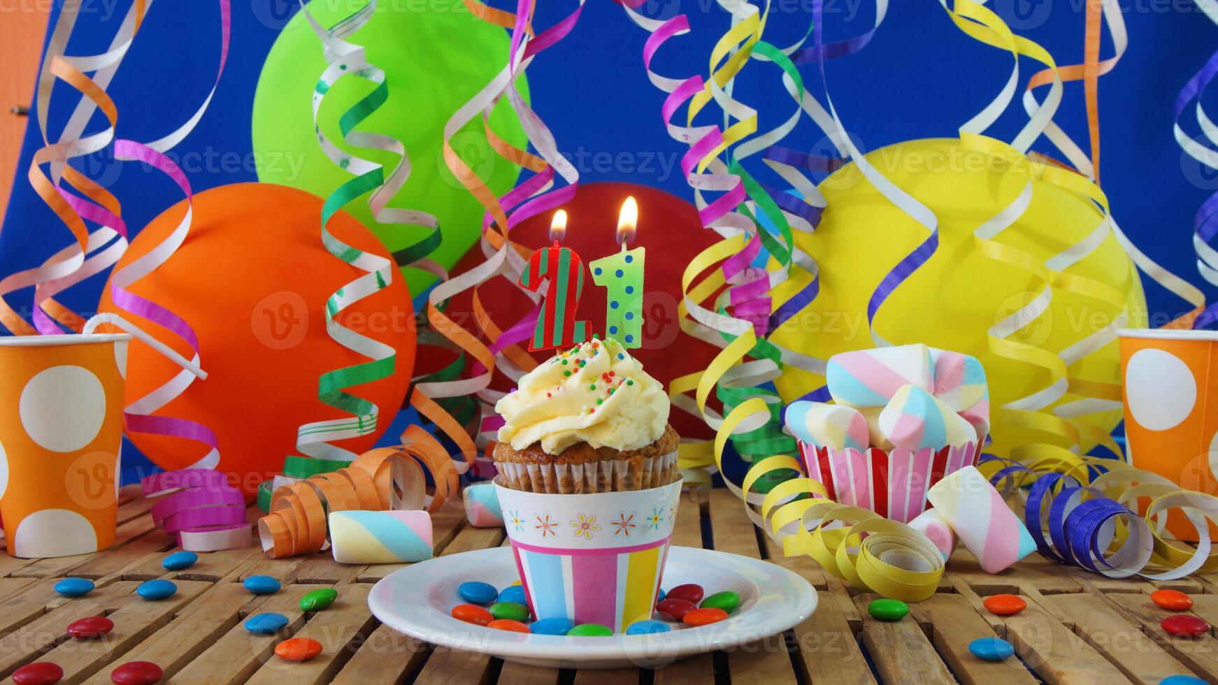 Birthday 21 cupcake with candles burning on rustic wooden table with background of colorful balloons, plastic cups and candies with blue wall in the background photo