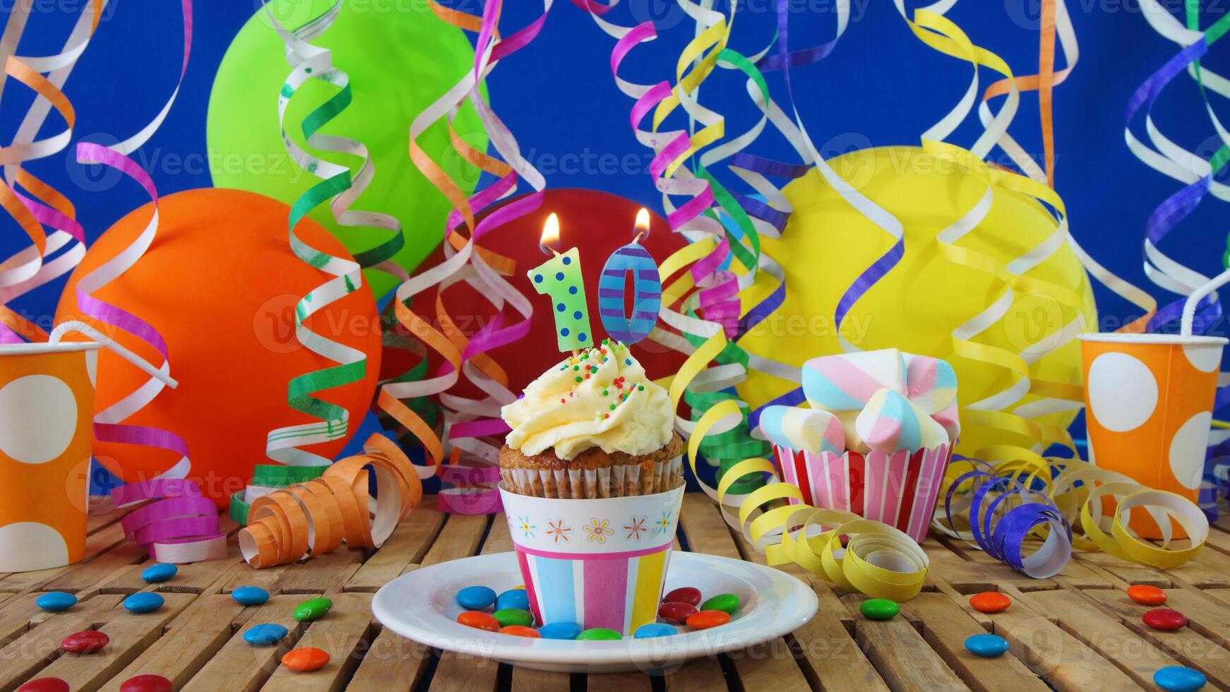 Birthday 10 cupcake with candles burning on rustic wooden table with background of colorful balloons, plastic cups and candies with blue wall in the background photo
