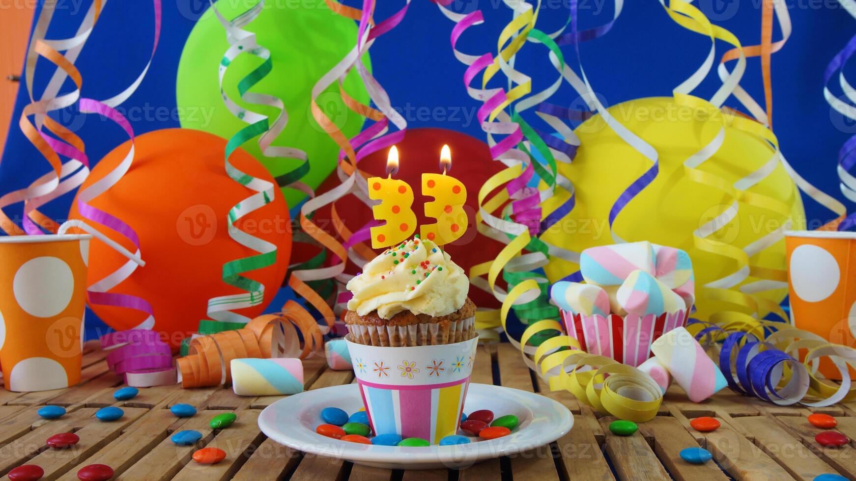 Birthday 33 cupcake with candles burning on rustic wooden table with background of colorful balloons, plastic cups and candies with blue wall in the background photo