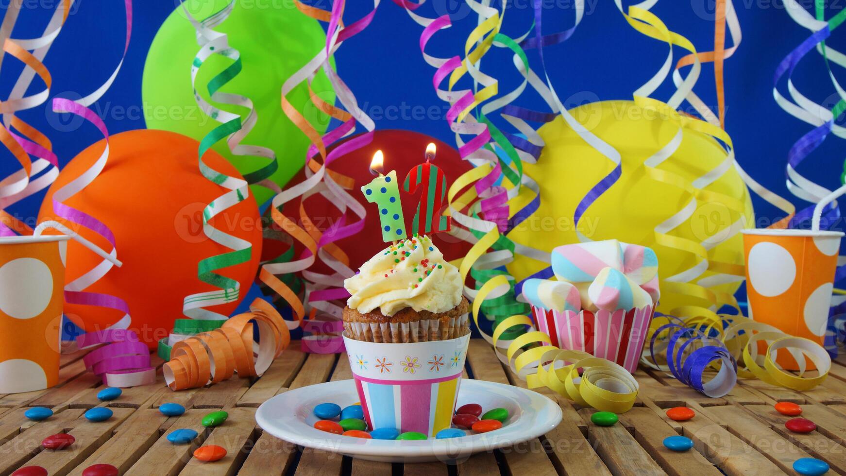 Birthday 12 cupcake with candles burning on rustic wooden table with background of colorful balloons, plastic cups and candies with blue wall in the background photo
