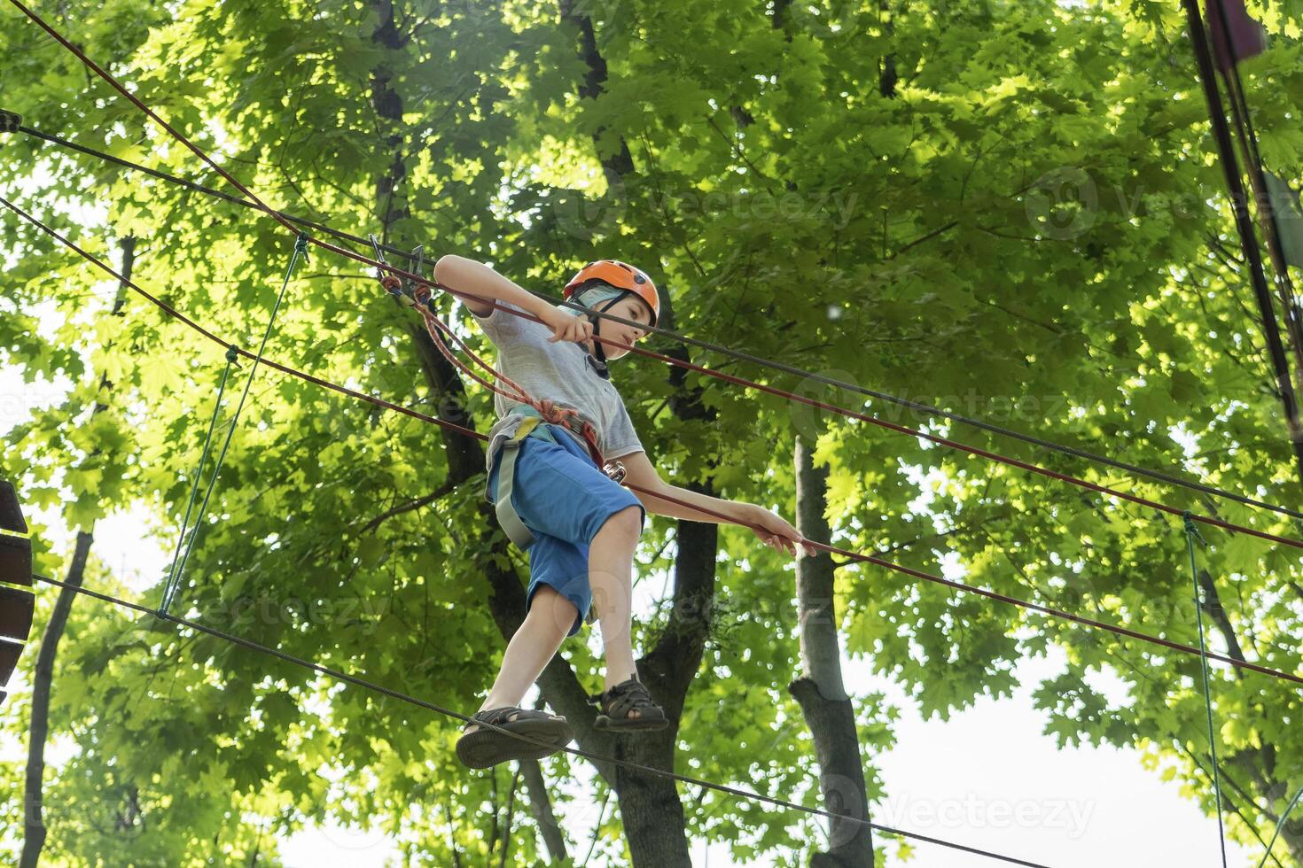 Rope park. A boy teenager in a helmet walks on suspended rope ladders. Carabiners and safety straps. Safety. Summer activity. Sport. Children's playground in nature in the forest photo