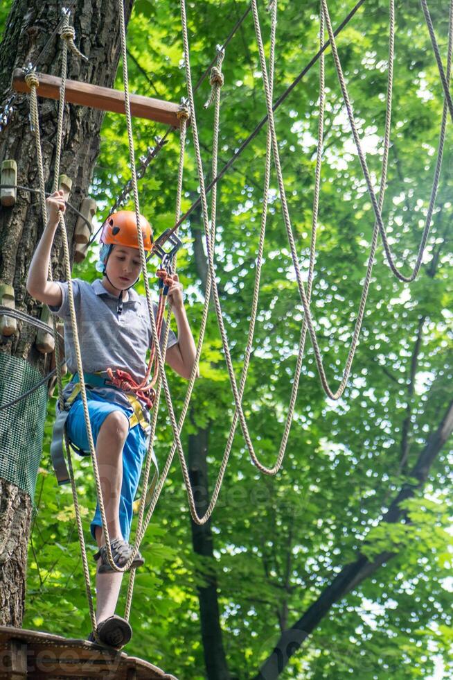 Rope park. A boy teenager in a helmet walks on suspended rope ladders. Carabiners and safety straps. Safety. Summer activity. Sport. Children's playground in nature in the forest photo
