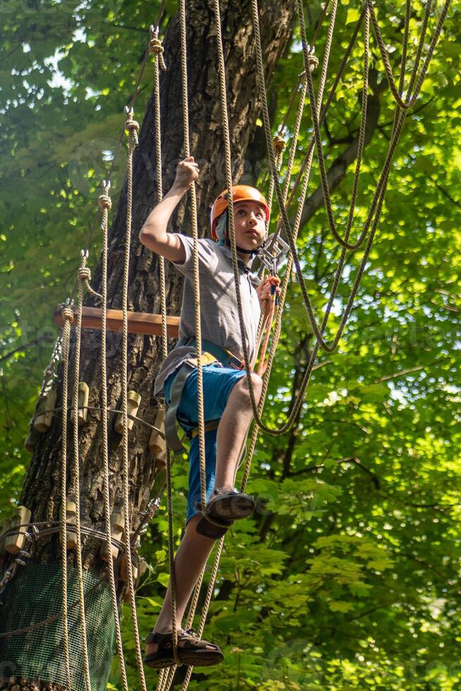 Rope park. A boy teenager in a helmet walks on suspended rope ladders. Carabiners and safety straps. Safety. Summer activity. Sport. Children's playground in nature in the forest photo