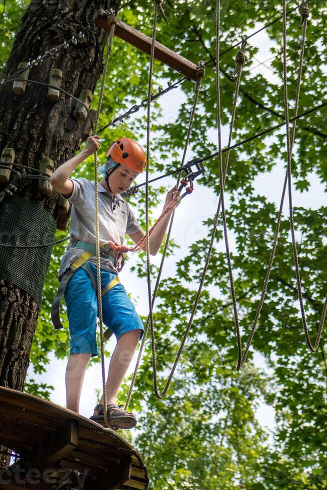 Rope park. A boy teenager in a helmet walks on suspended rope ladders. Carabiners and safety straps. Safety. Summer activity. Sport. Children's playground in nature in the forest photo