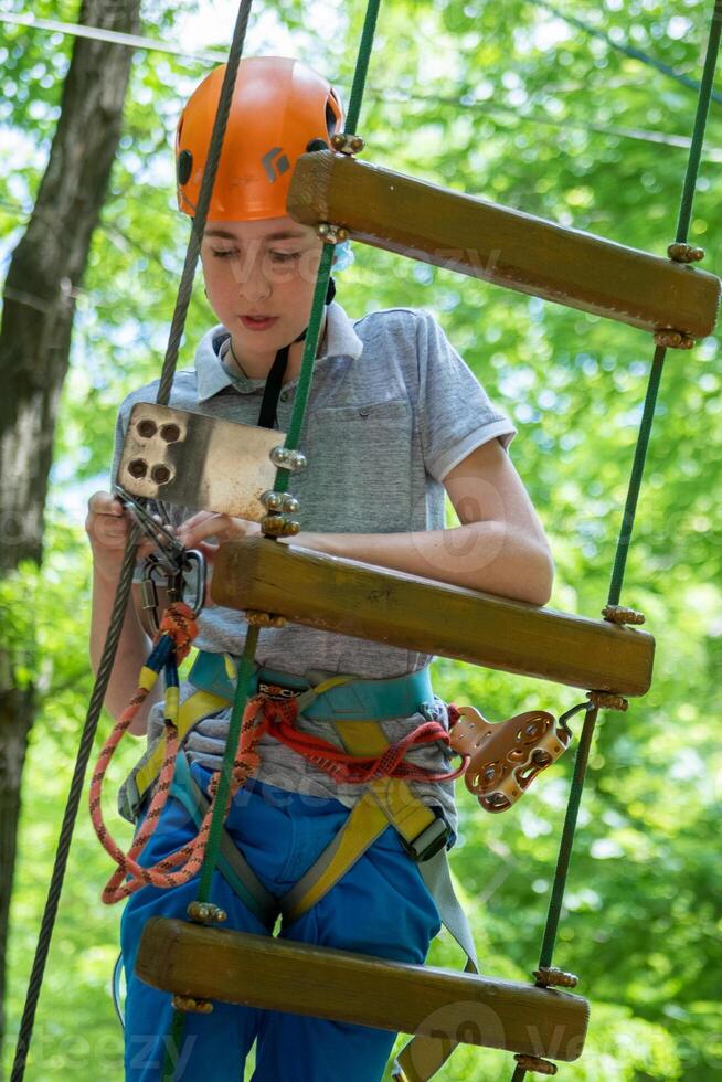 Rope park. A boy teenager in a helmet walks on suspended rope ladders. Carabiners and safety straps. Safety. Summer activity. Sport. Children's playground in nature in the forest photo