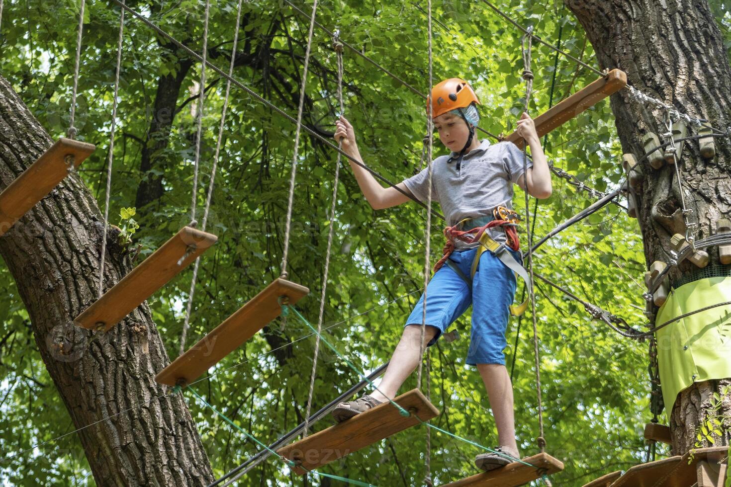 Rope park. A boy teenager in a helmet walks on suspended rope ladders. Carabiners and safety straps. Safety. Summer activity. Sport. Children's playground in nature in the forest photo