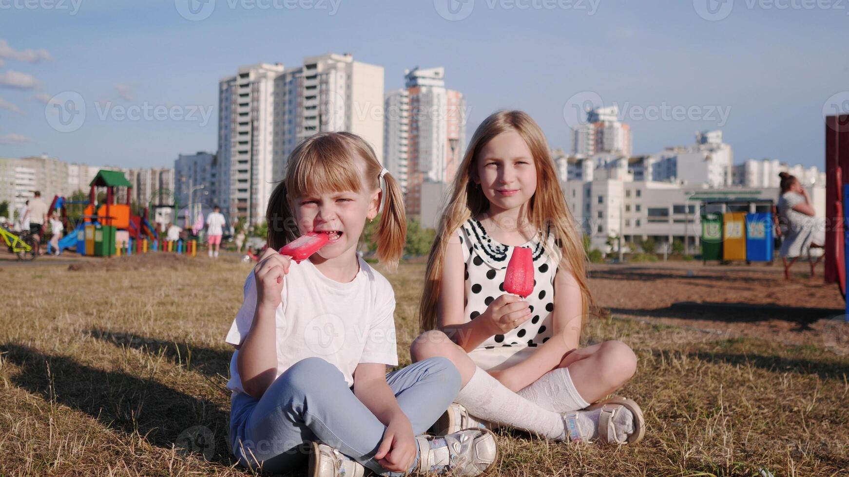 Two little girls sisters sit on the lawn in the city in the summer and eat delicious ice cream against the blue sky photo