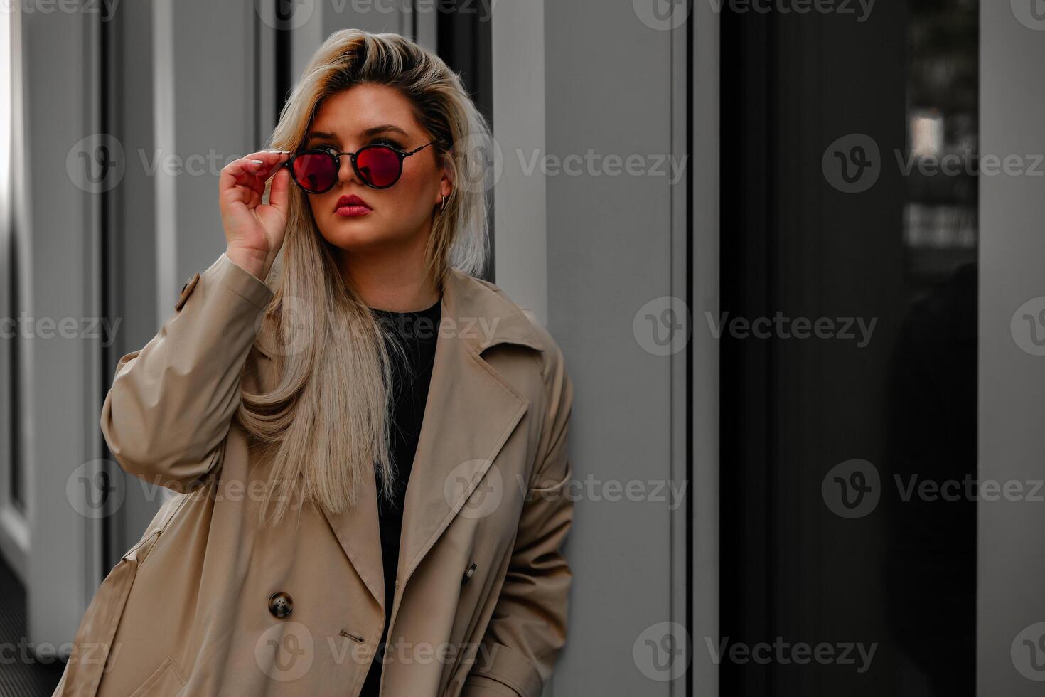 woman in a tan trench coat with a black shirt and red sunglasses in the city photo