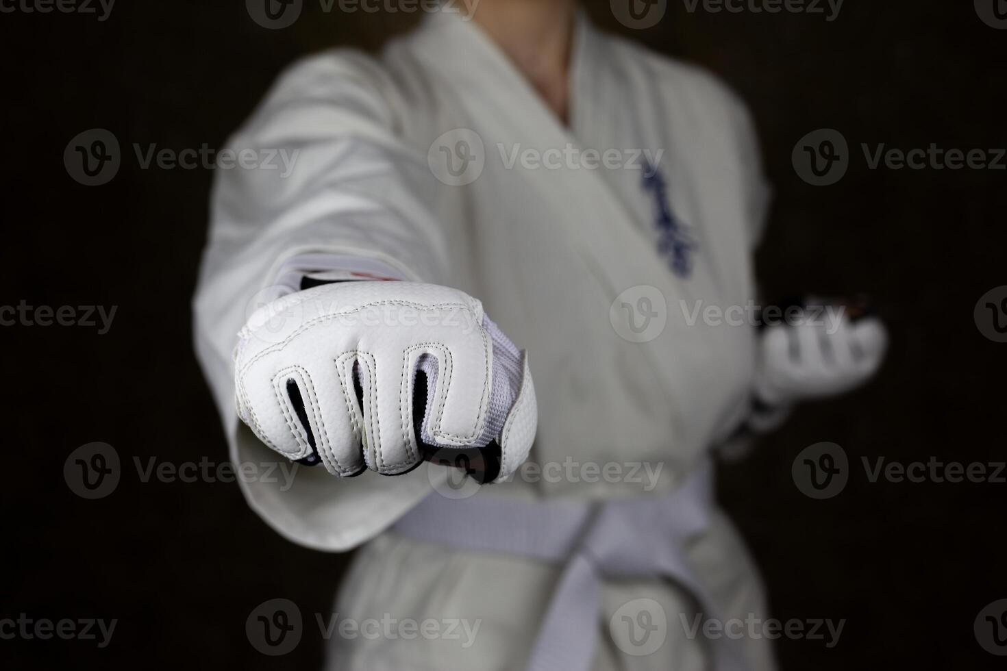 Young woman in kimono and white belt throws punch in protective gloves in Kyokushin karate to camera photo