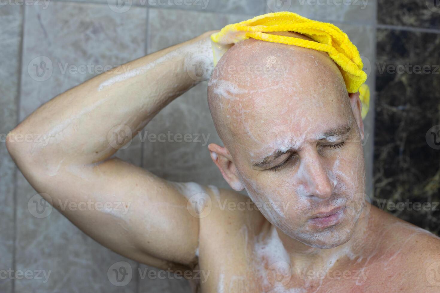 Man with closed eyes in shower washes his bald hairless head with washcloth and foam photo