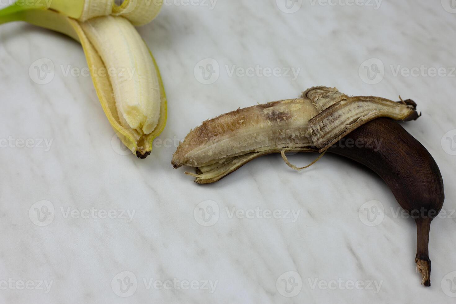Rotten spoiled banana in open peel next to fresh banana fruit on white table photo