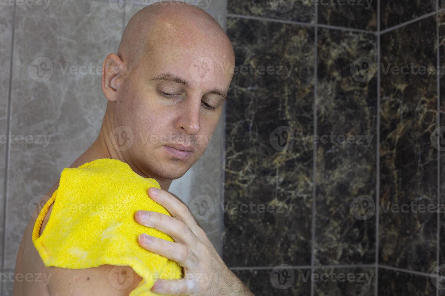 Man without hair washes himself in shower with washcloth, bald guy takes care of his skin in the shower with a special product with foam photo