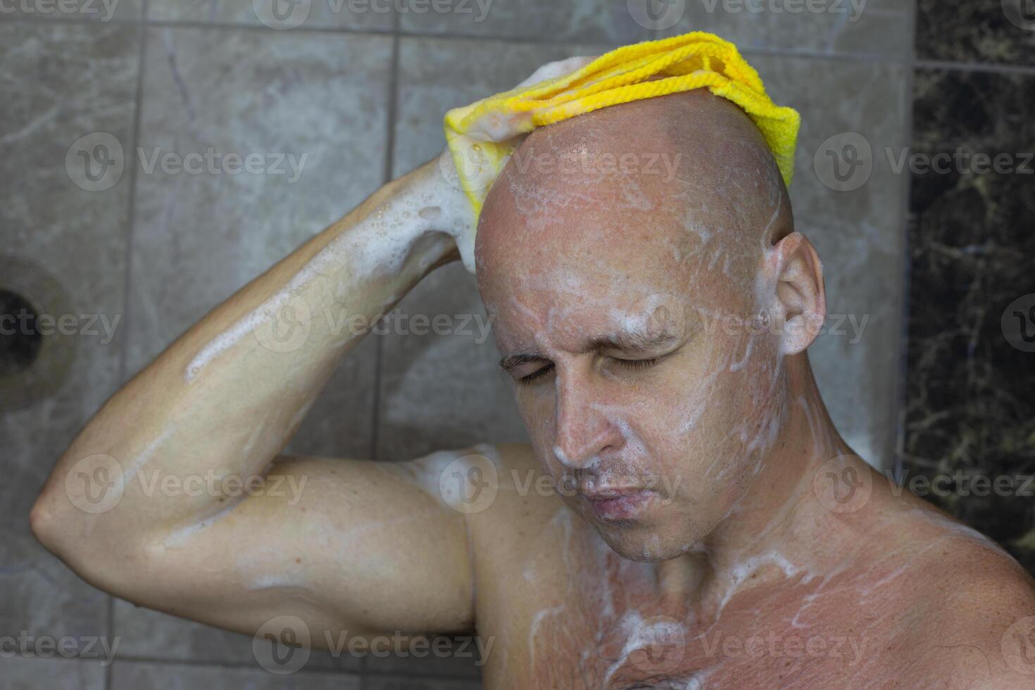 Bald European man washes his head in the shower with foam and washcloth, taking care of the cleanliness of his body in the shower photo