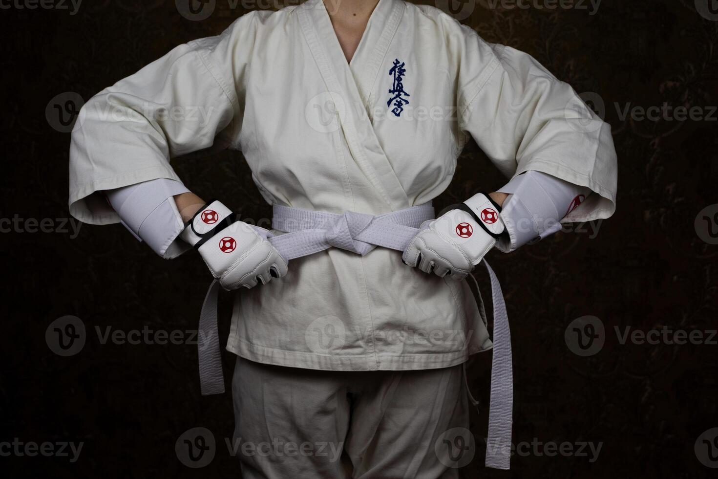 Girl Kyokushin karate athlete in kimono and protective gloves tightens white belt around her waist. Inscription in Japanese. Kyokushinkai photo